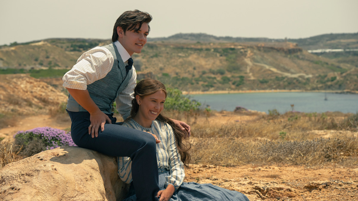 Millie Bobby Brown as Enola Holmes and Louis Partridge as Lord Tewkesbury sitting on rocks by the sea in Enola Holmes 3