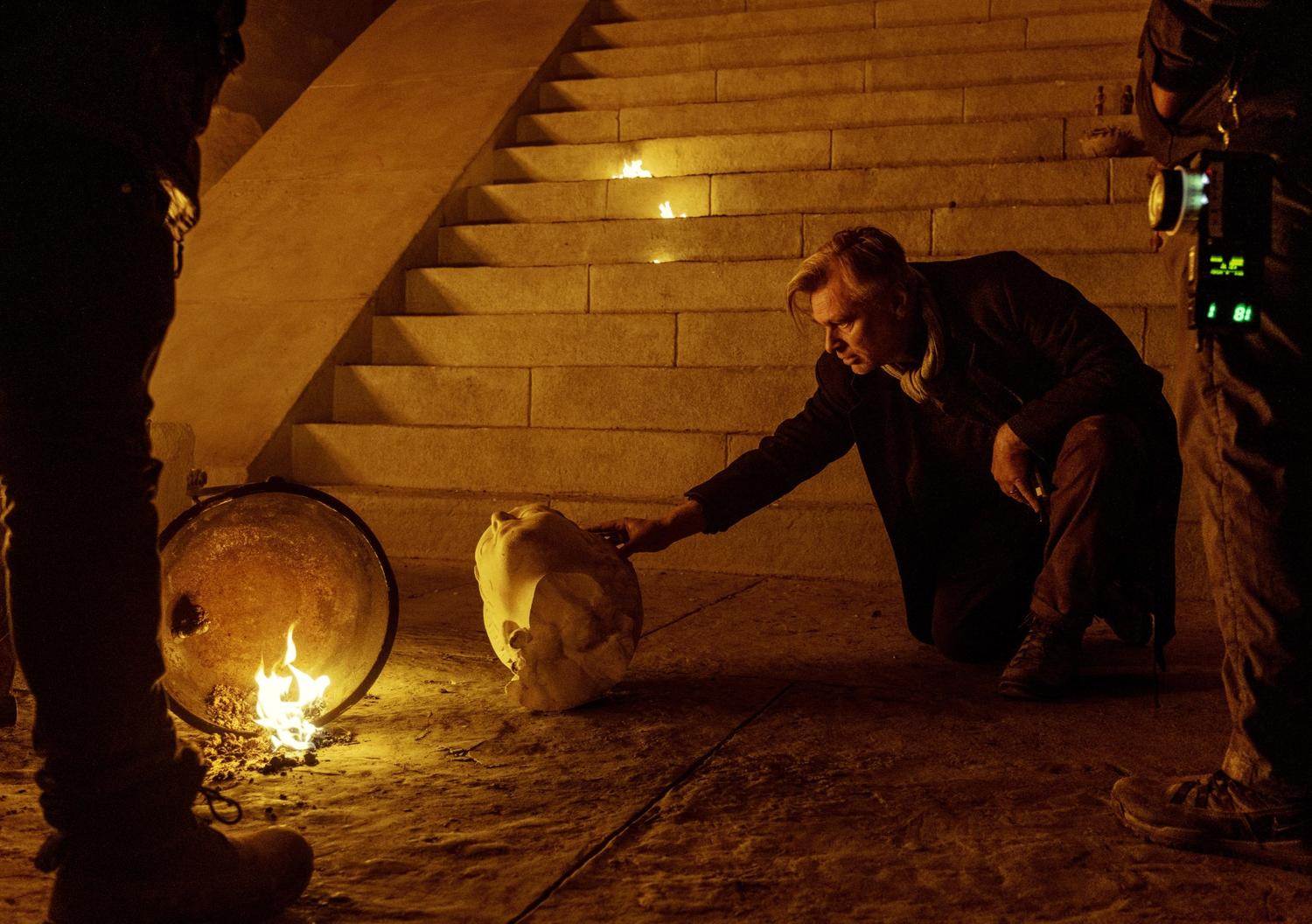 Behind-the-scenes photo from The Odyssey showing the director examining a damaged statue head beside practical fire effects